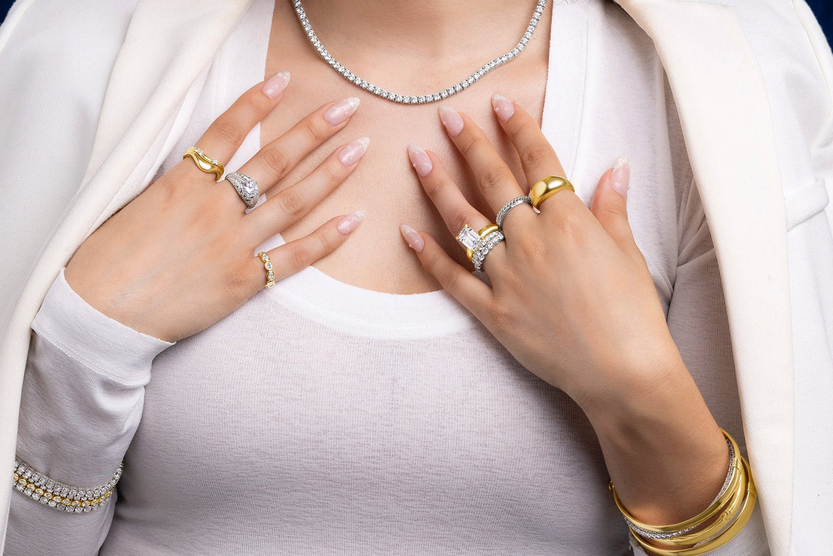 A close-up of a woman wearing multiple gold rings with diamond accents, a diamond necklace, and a diamond bracelet, styled with a white blazer and white top against a dark blue velvet curtain.