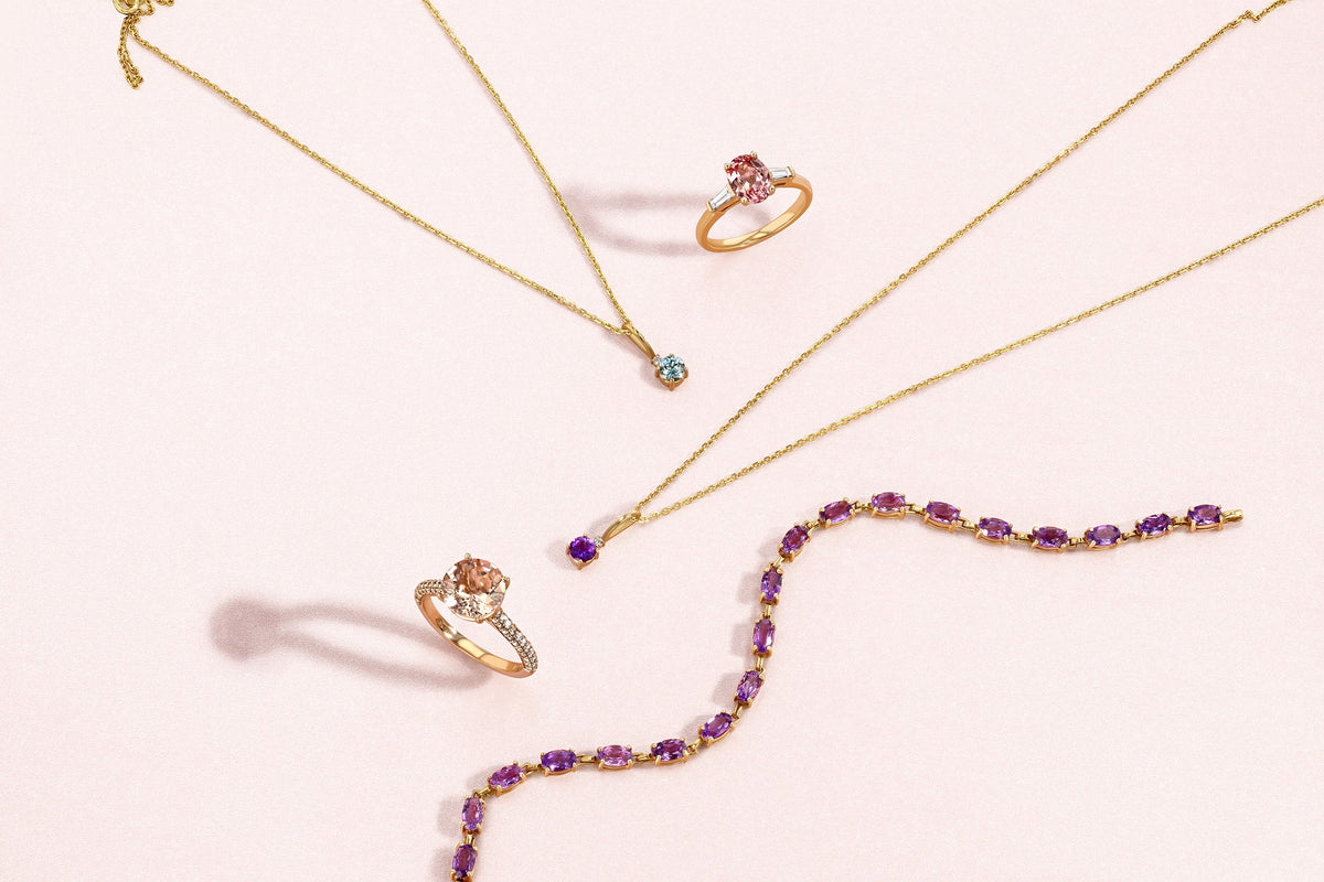 Two gold rings, two gold pendant necklaces, and a purple gemstone bracelet arranged on a pale pink surface.