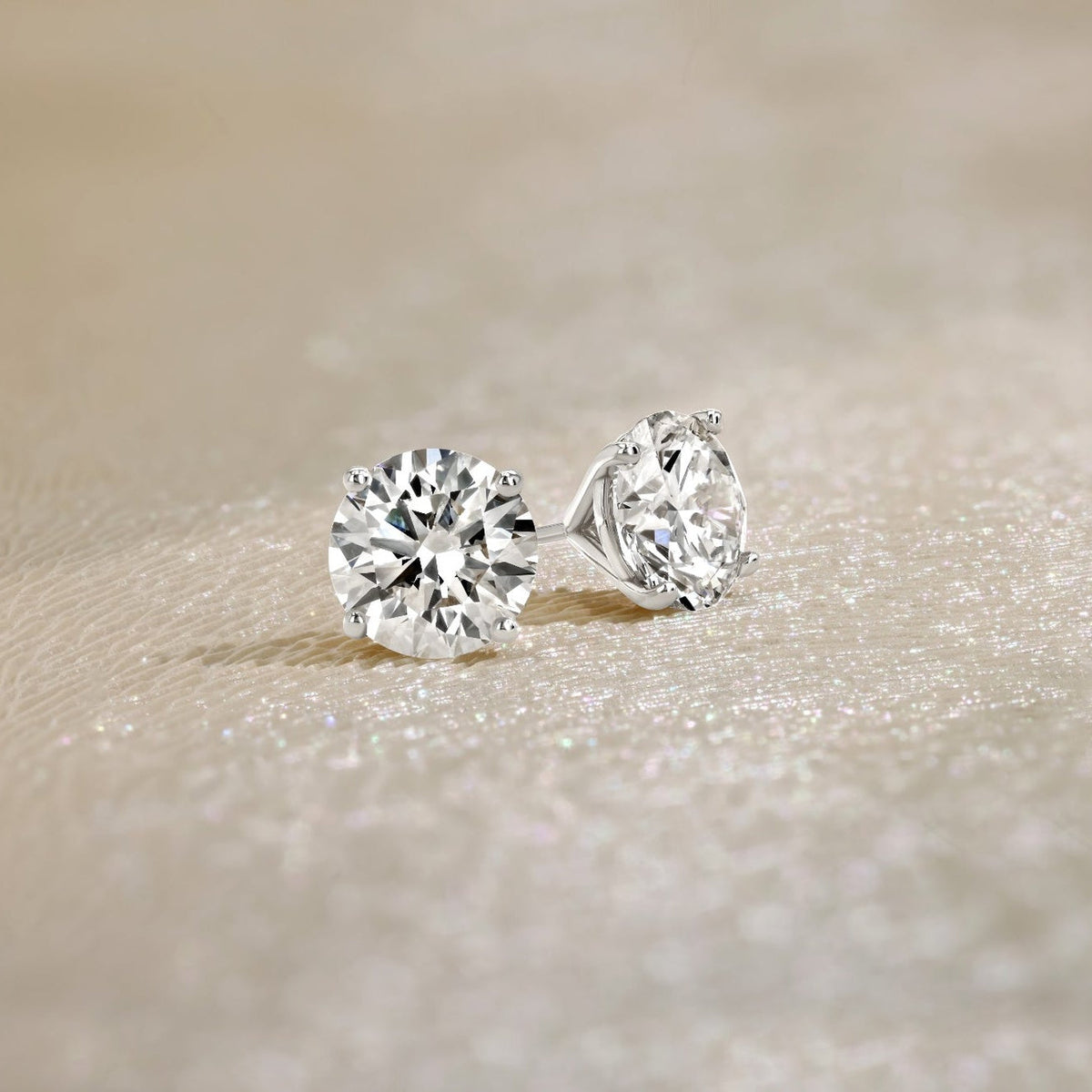 A pair of round-cut diamond stud earrings in a silver-toned setting, resting on a textured beige surface.
