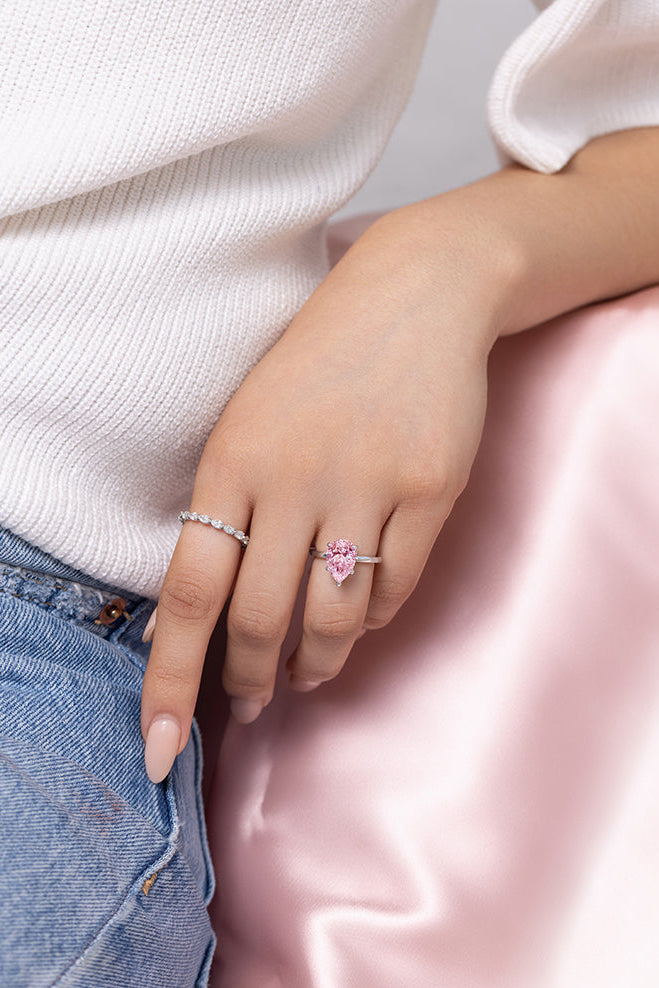 A close-up lifestyle shot of a person's hand resting on a pink satin surface, wearing a silver band ring with a pink pear-shaped diamond and a thin silver spacer ring.