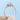 A Granada round halo engraved engagement ring held between two fingers against a light blue background, featuring a round center diamond and intricate engraving along the band.
