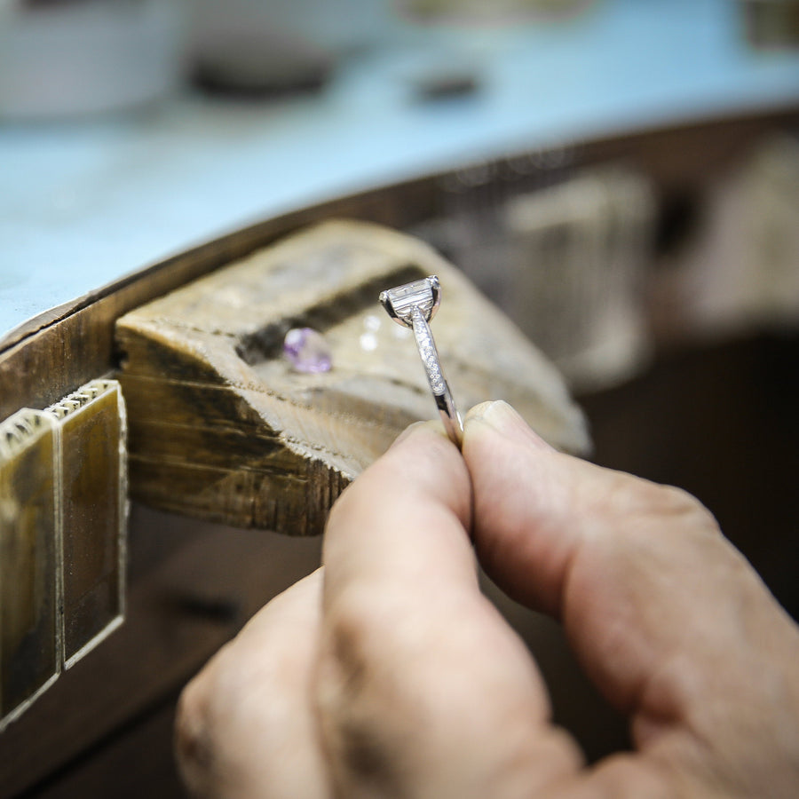 A close-up of a jeweler's hands holding a small silver ring on a wooden ring press inside a workshop.