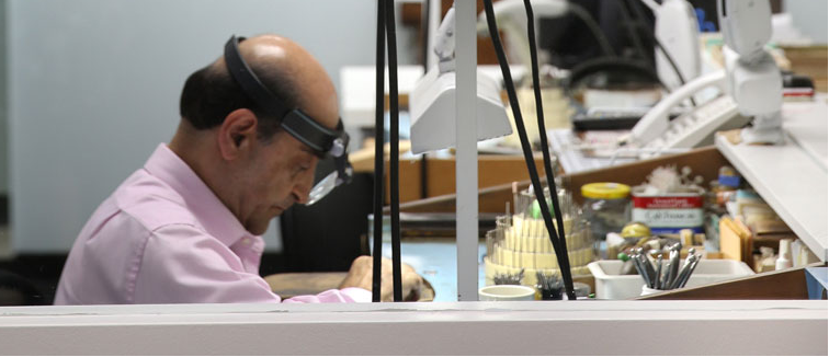 A man wearing a pink shirt and a black magnifying headlamp sits at a workbench in a workshop or office setting with tools and equipment in the background.
